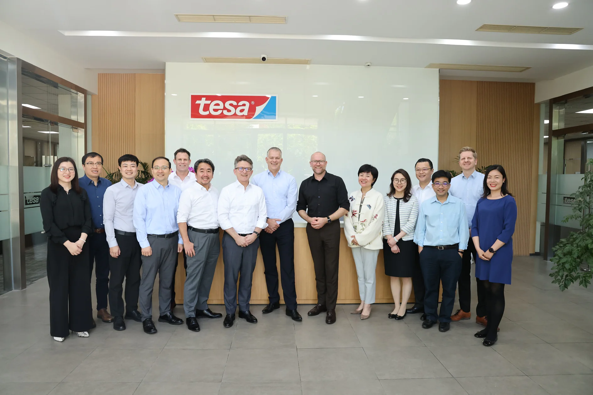 A group of 16 people, composed of men and women in business attire, stand in a well-lit office lobby. They are posing in front of a wall with a sign that reads tesa tape. Most of them are smiling. The floor is tiled, and there are glass doors on both sides. (This text has been generated by AI)