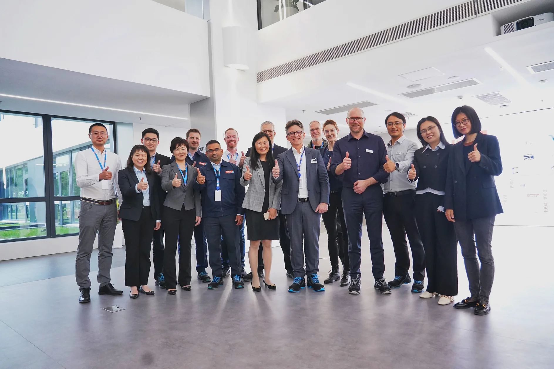 A group of people in business attire stands together in a bright office space, all giving thumbs up gestures. They appear to be participating in a team meeting or corporate event. Large windows are visible on the left side of the room. (This text has been generated by AI)