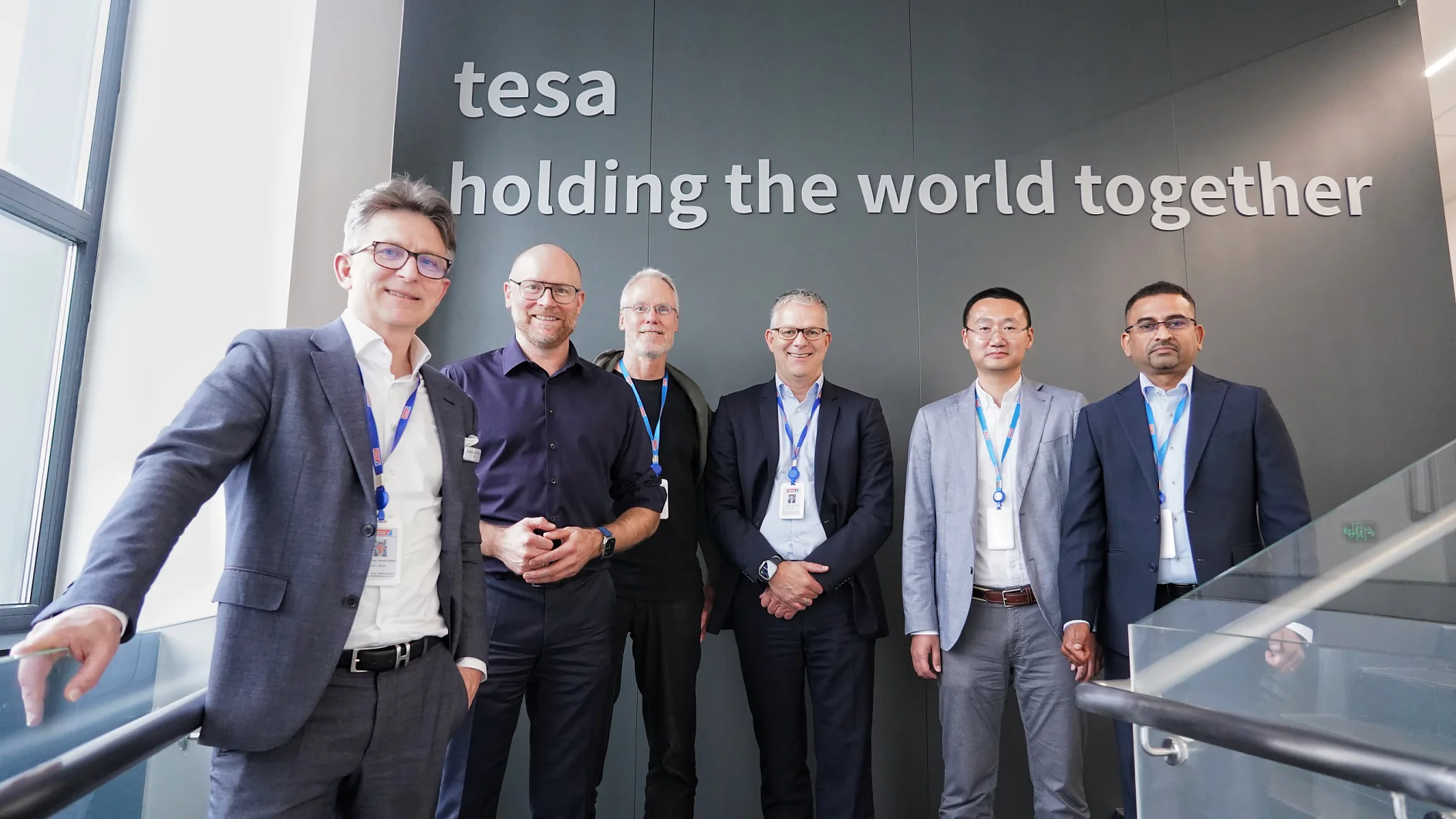 Six men in business attire stand in a line, smiling at the camera. They are in an office environment with a white wall behind them displaying the text tesa tape holding the world together. Some wear ID badges on lanyards. (This text has been generated by AI)