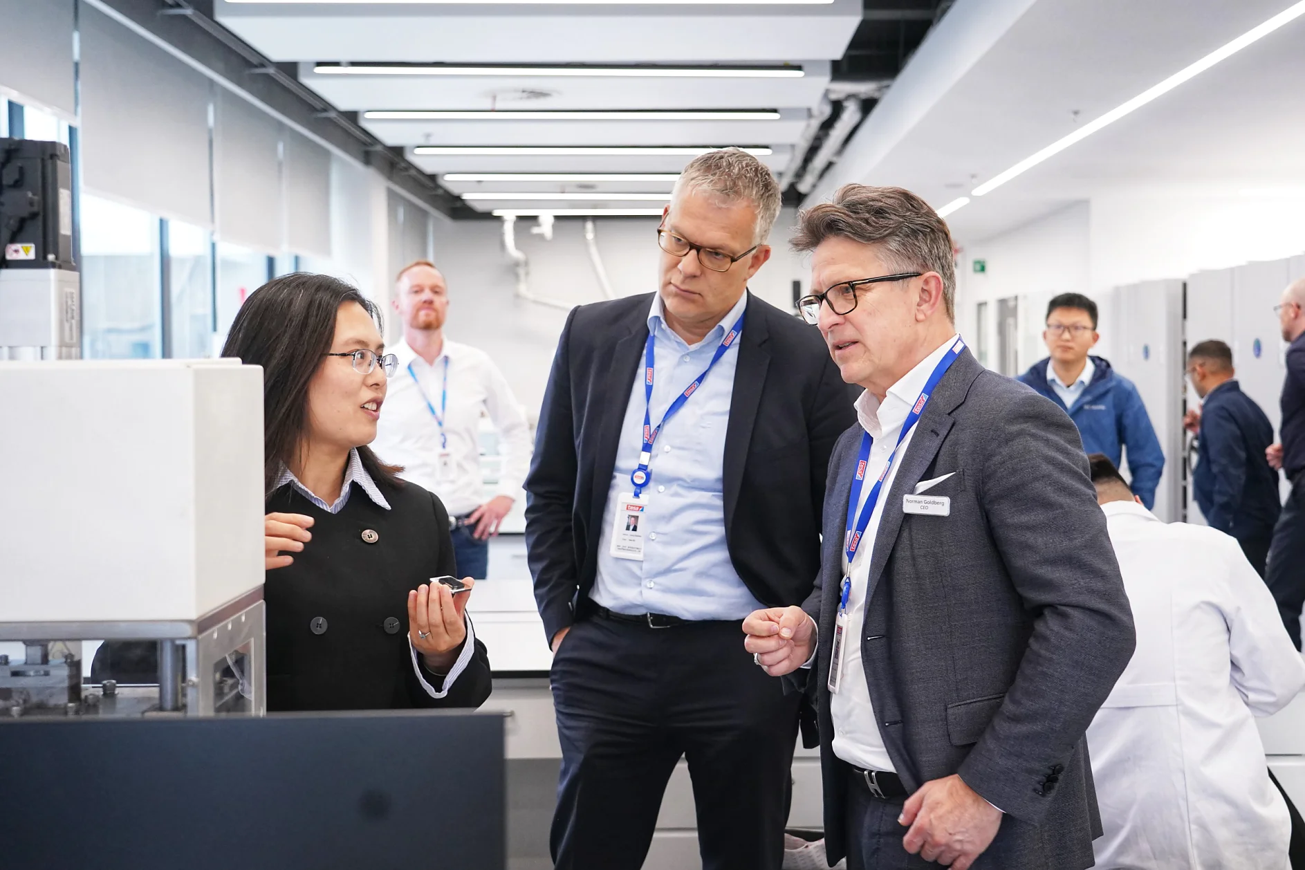 A woman is explaining something to two men in suits in a modern office space, using tesa tape. Other people are in the background, and everyone is wearing ID badges. The setting appears to be a professional or corporate environment where tesa tape is frequently utilized. (This text has been generated by AI)