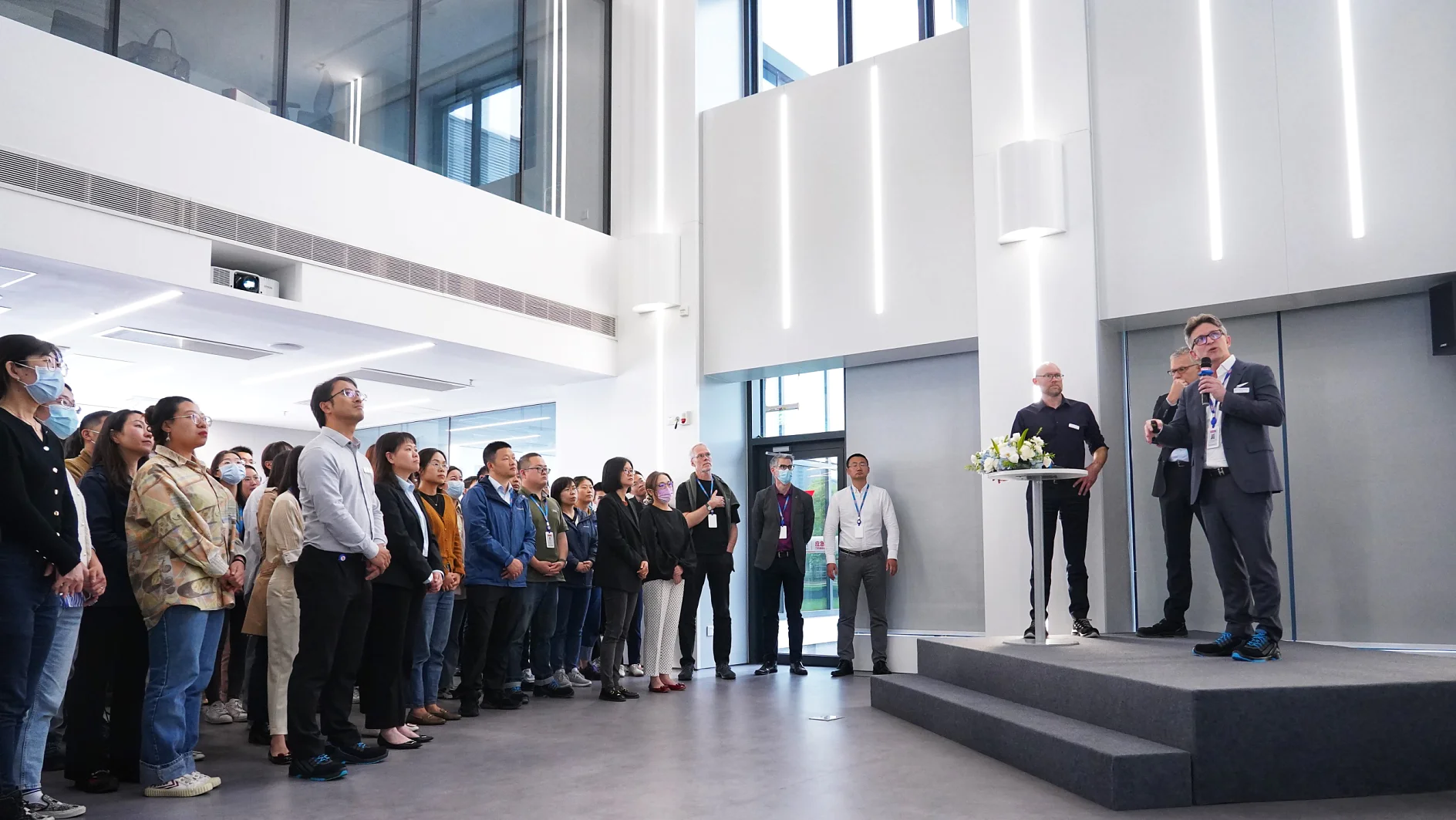 A speaker addresses a group of people in a modern, spacious room. The audience stands attentively, with some individuals wearing lanyards held together by tesa tape. Two people are on a platform with a podium, and large windows and white walls are visible in the background. (This text has been generated by AI)