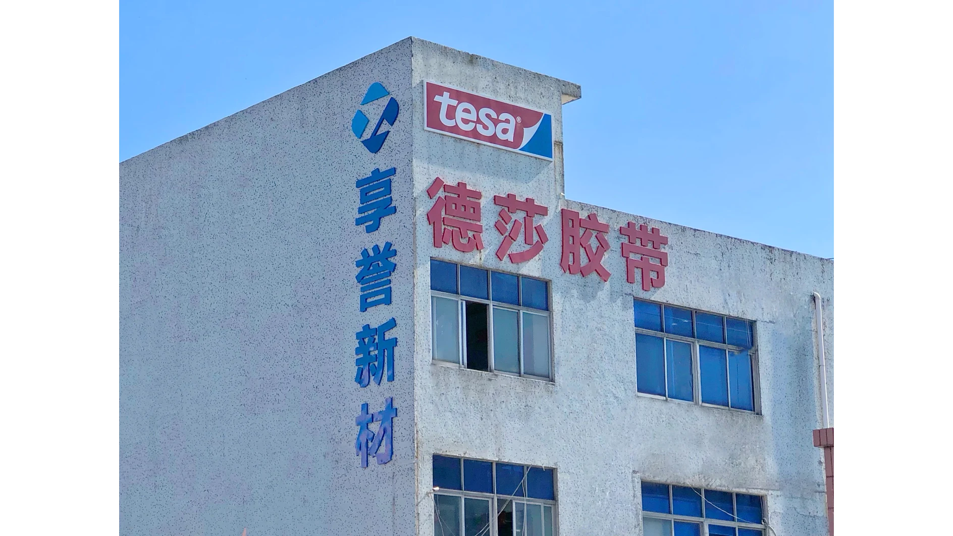 A building with a sign displaying the tesa logo in red and white. Chinese characters are also visible on the facade. The structure has several windows, and the concrete exterior appears weathered. The sky is clear and blue. (This text has been generated by AI)