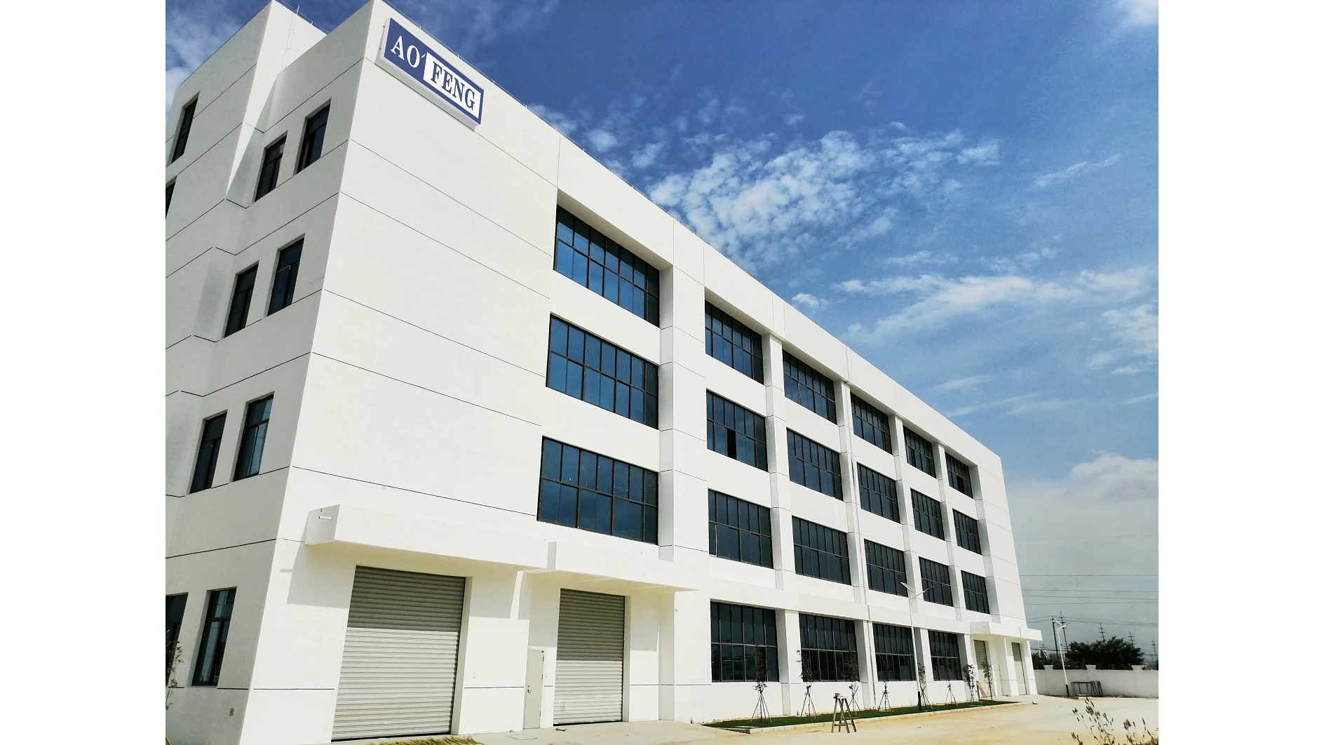 A four-story white industrial building with large blue-tinted windows is shown under a partly cloudy sky. Two large garage doors are visible on the ground floor. The sign AO FENG is mounted on the buildings corner, held securely in place by tesa tape. (This text has been generated by AI)