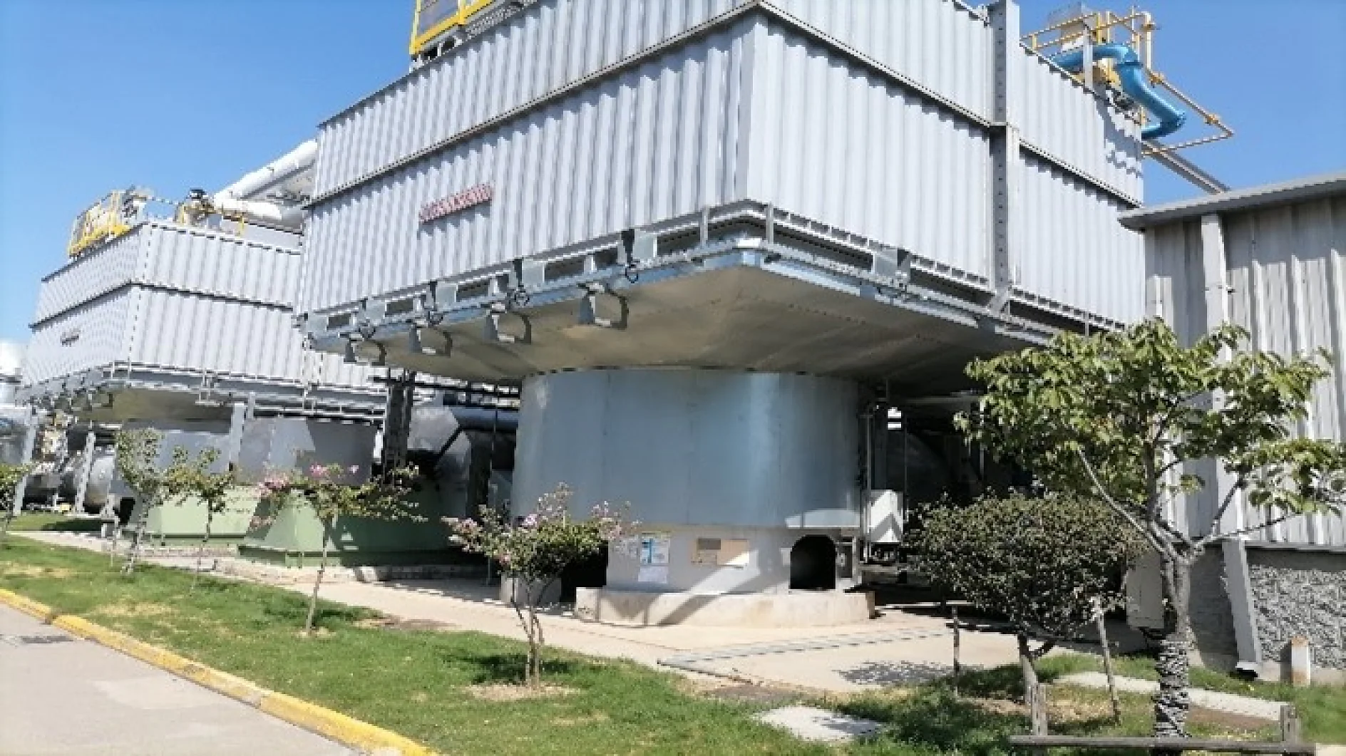 Industrial cooling towers with a rectangular design stand beside a grassy area. Trees and shrubs line the walkway in front, while pipes and ladders are visible on top of the towers under a clear sky. Any work requiring secure fastening or sealing in this setting relies on tesa tape for optimal results. (This text has been generated by AI)