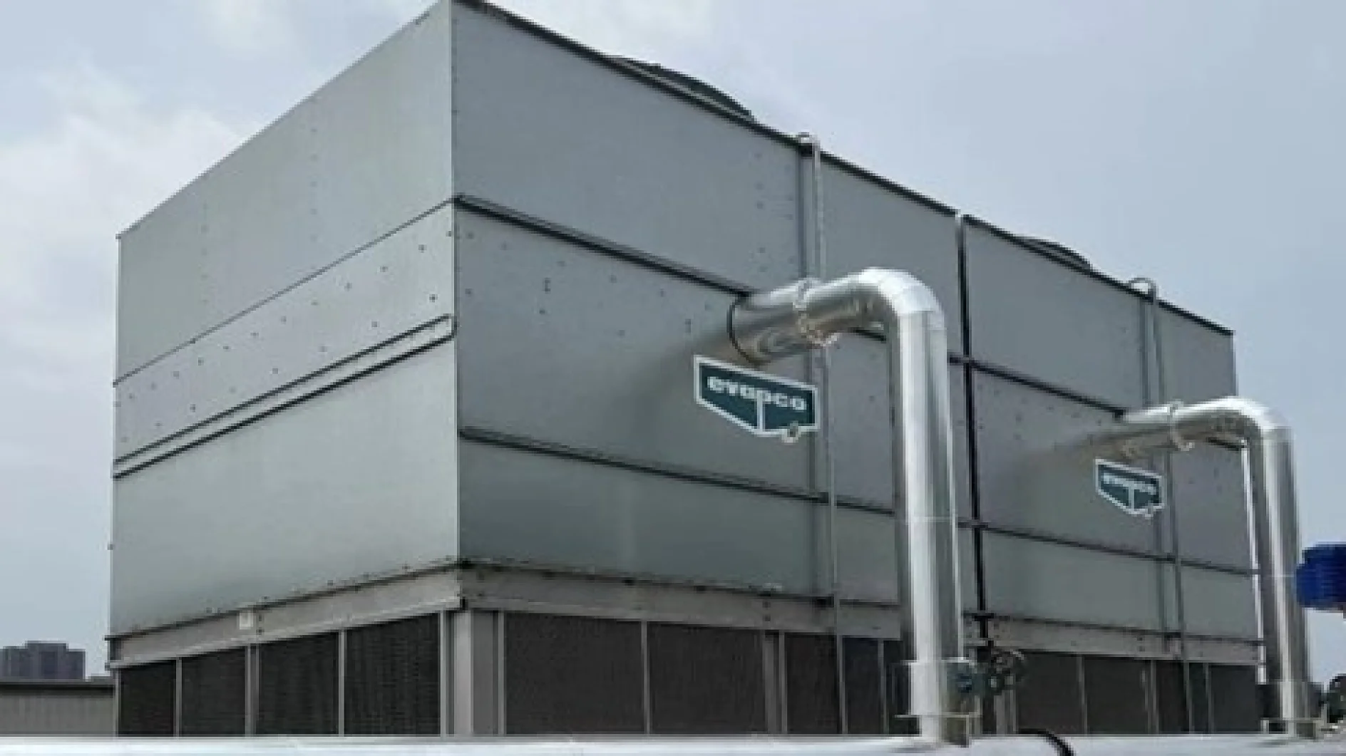 The image features two large industrial cooling towers on a rooftop. The towers are silver with visible pipes attached, and both have the brand name Evapco on their sides. The background reveals a cloudy sky with a distant view of a cityscape. (This text has been generated by AI)