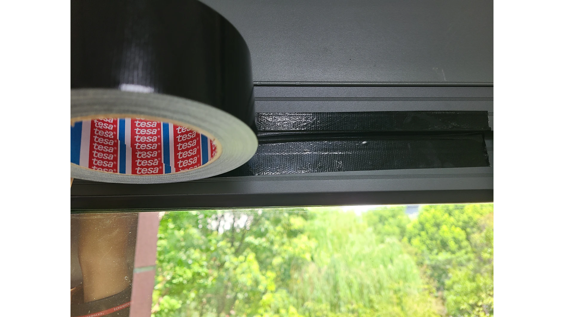 A roll of tesa tape-branded black tape is partially unrolled above a window frame. The tesa tape has been applied in two parallel strips on the frame. Outside the window, greenery and part of a red brick wall are visible. (This text has been generated by AI)