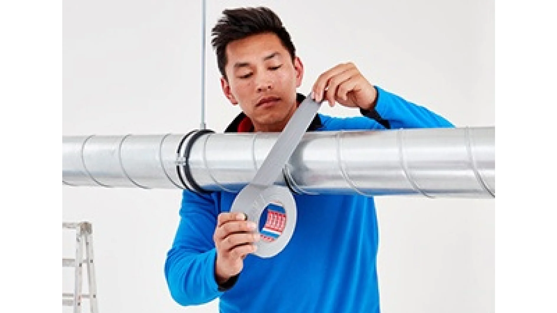 A person wearing a blue jacket is applying tesa tape to a metal pipe. A ladder is partially visible on the left side. The background is plain white. (This text has been generated by AI)