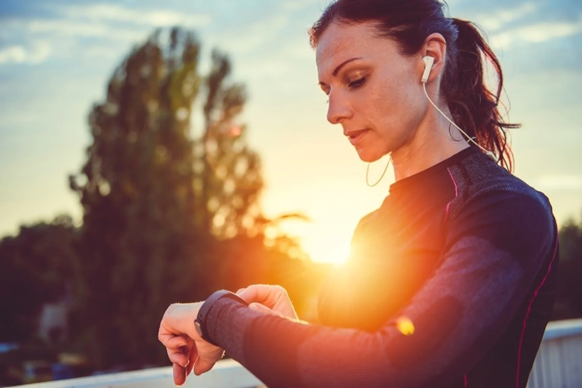 A woman in athletic wear, with earphones in, stands outdoors at sunset. She is looking at a smartwatch on her wrist. A tree is blurred in the background, and the setting sun creates a warm glow. (This text has been generated by AI)