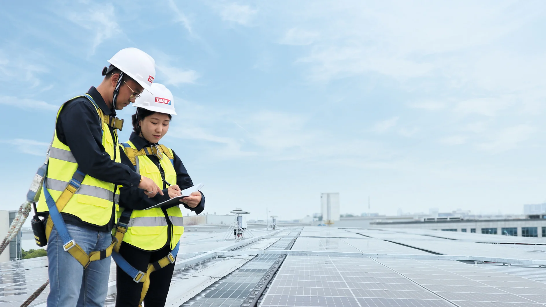 Two people in safety gear stand on a rooftop covered with solar panels. They wear yellow high-visibility vests, hard hats, and safety harnesses. One person holds a document, and they appear to be discussing it. The sky is clear and blue. (This text has been generated by AI)