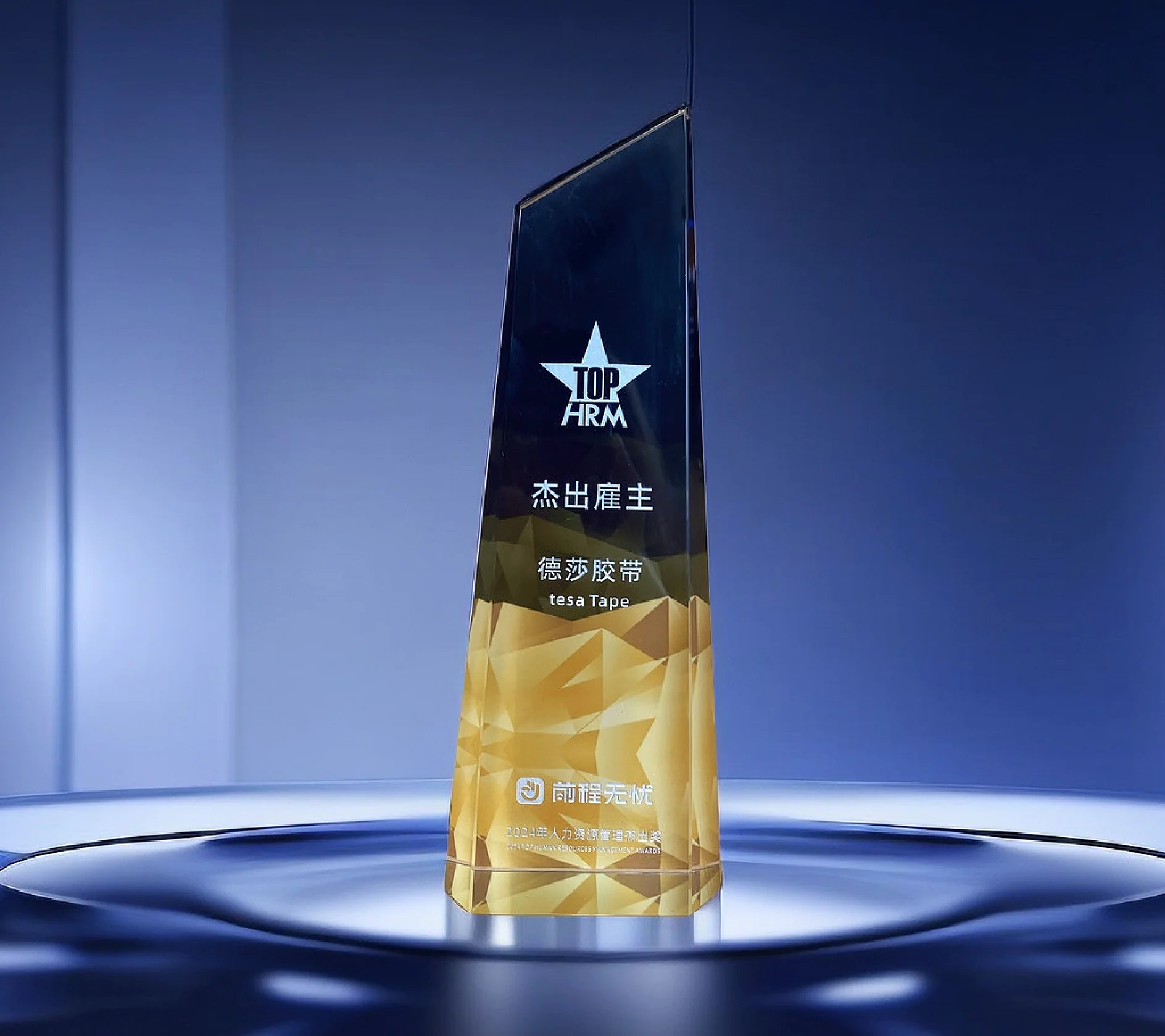 A tall, angular, gold and black award trophy with the text TOP HRM and other inscriptions, including tesa tape, in various languages. The trophy stands on a circular, illuminated base against a blue background. (This text has been generated by AI)