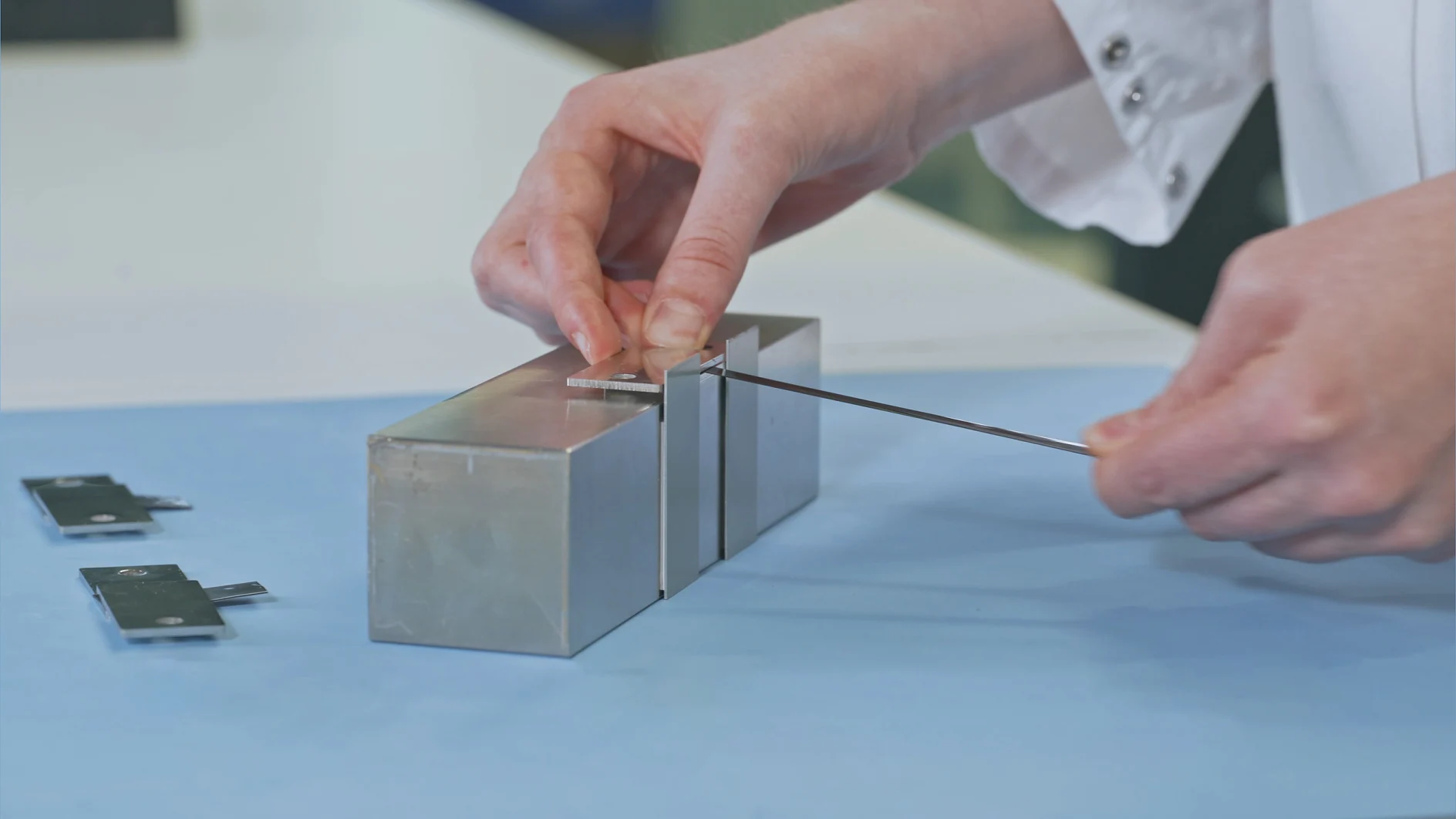 A person in a lab coat is adjusting a metallic test sample on a block using a gauge. The setup is on a blue work surface, and additional samples are placed nearby, secured with tesa tape. (This text has been generated by AI)