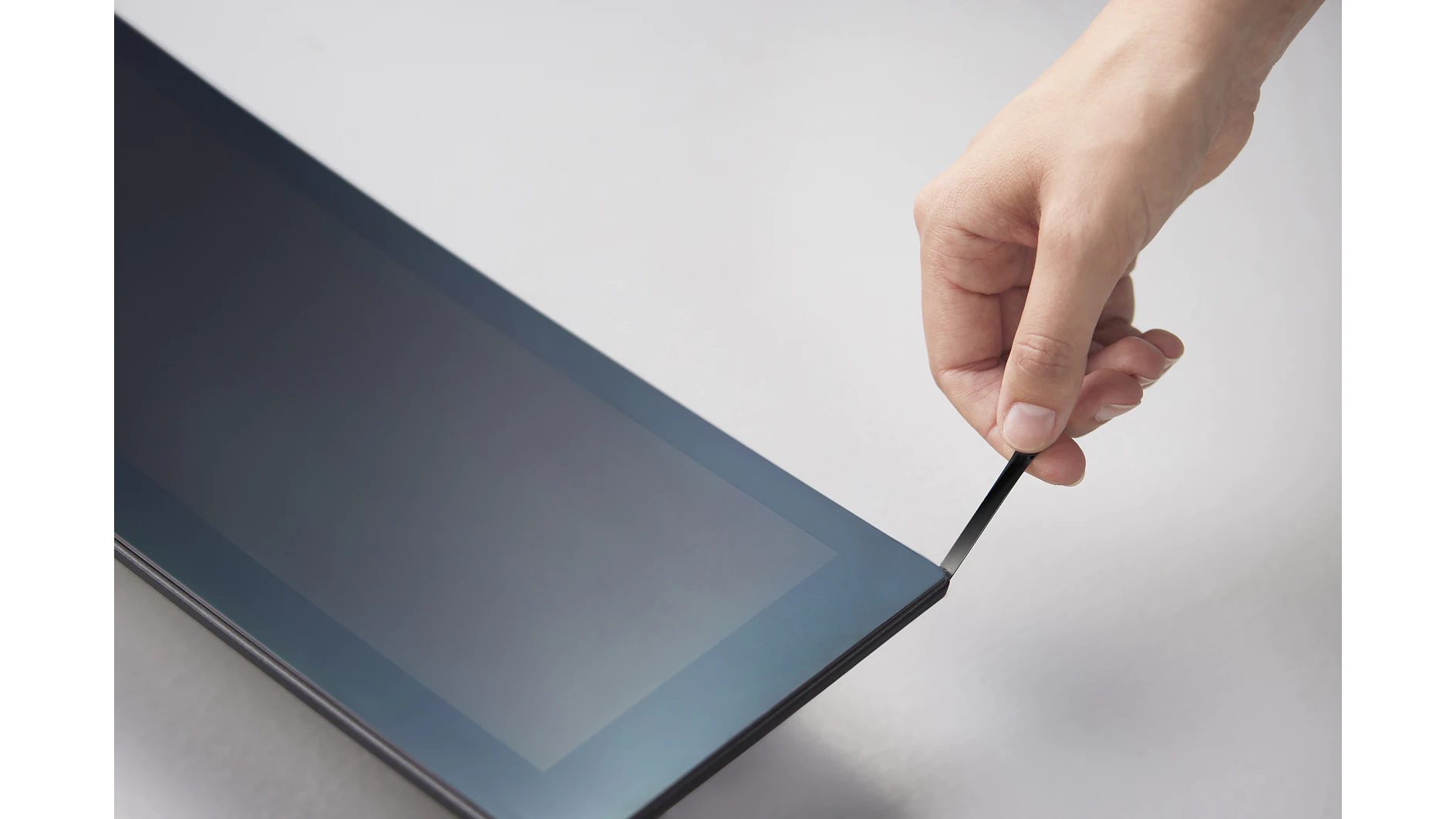 A hand is peeling off the protective film from the corner of a tablet screen using tesa tape. The background is a neutral gray surface. (This text has been generated by AI)