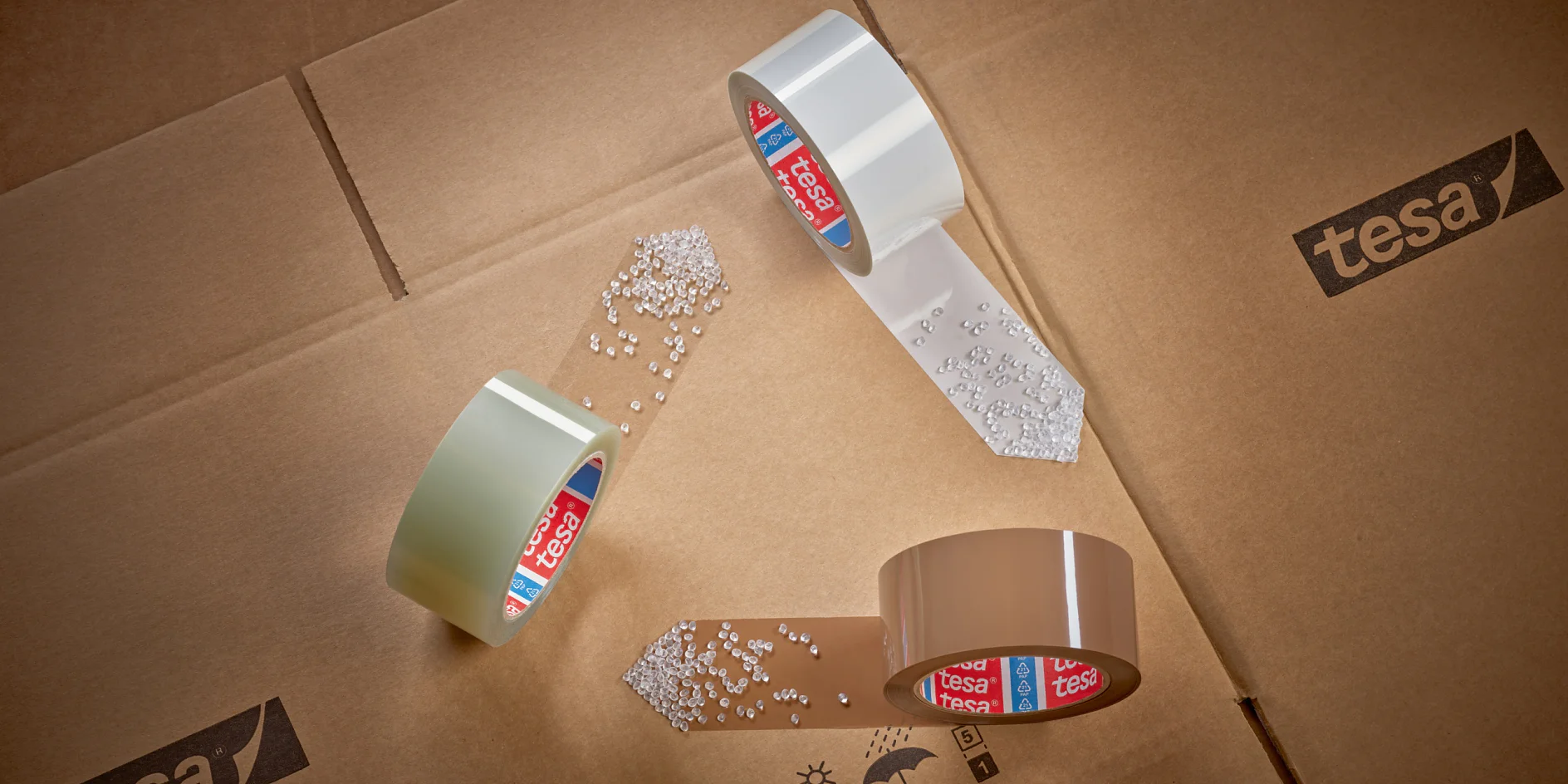 Three rolls of tesa tape are arranged in a triangular formation on cardboard boxes. White pellets are scattered near the ends of the tesa tape. Tesa branding is visible on the tesa tape cores and the boxes. (This text has been generated by AI)