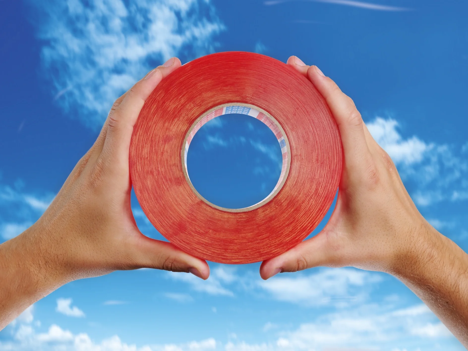 Two hands hold a large roll of red tesa tape against a backdrop of a blue sky with scattered clouds. The tesa tape roll forms a circular frame through which the sky is visible. (This text has been generated by AI)