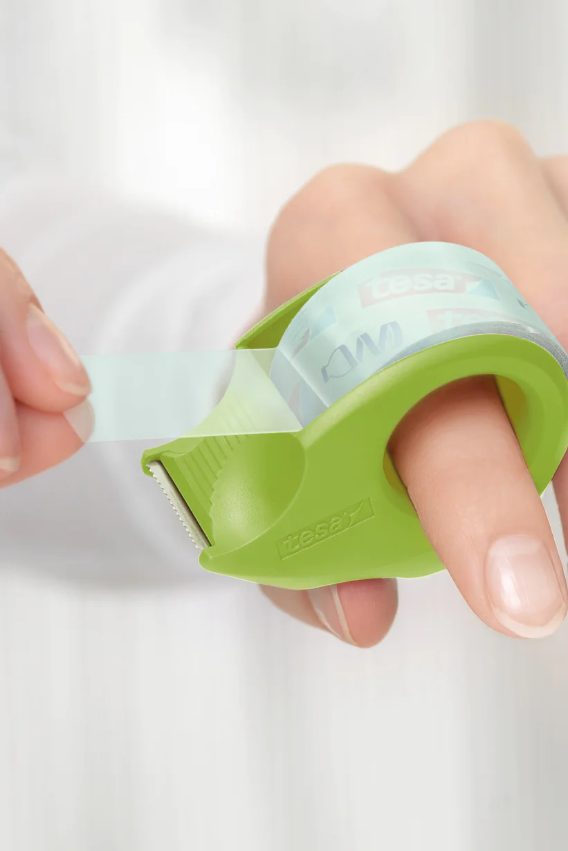 Close-up of a person dispensing clear adhesive tesa tape using a green handheld tape dispenser. The dispenser is wrapped around the index finger, and the person is pulling the tesa tape with the other hand. The background is softly blurred. (This text has been generated by AI)