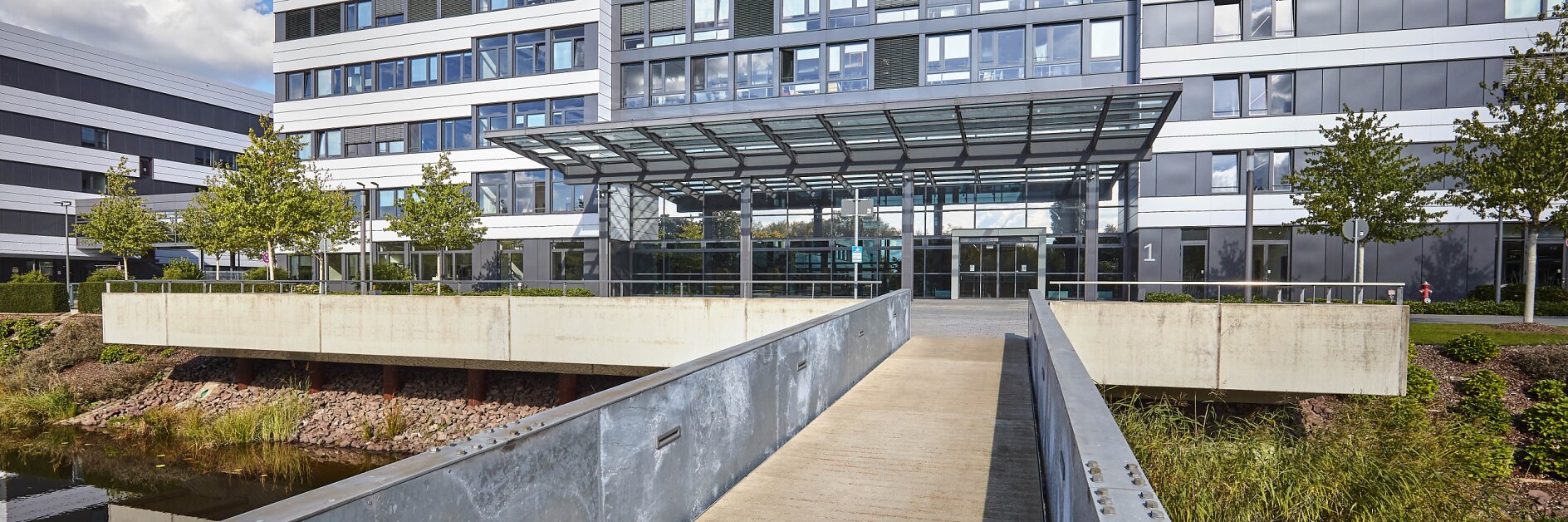 Exterior view of a modern office building with a glass and metal facade. A pedestrian bridge leads to the main entrance, featuring a glass canopy. The building is surrounded by greenery and water, and a tesa tape logo is visible at the top. (This text has been generated by AI)