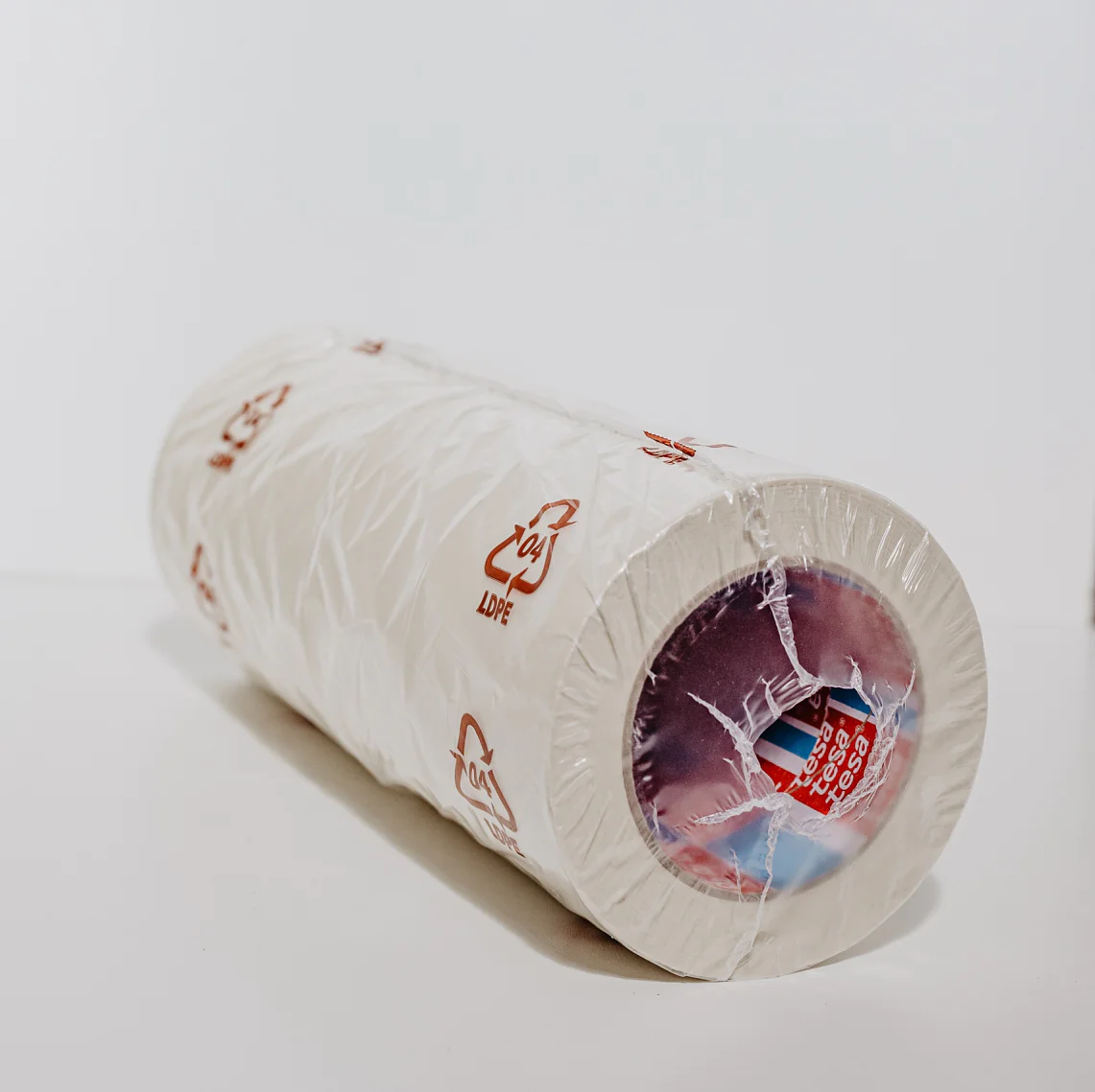 A roll of white plastic wrap is standing upright against a plain white background. The packaging is partially transparent, revealing the inner cardboard tube and multicolored label. Red recycling symbols and text can be seen on the plastic. (This text has been generated by AI)