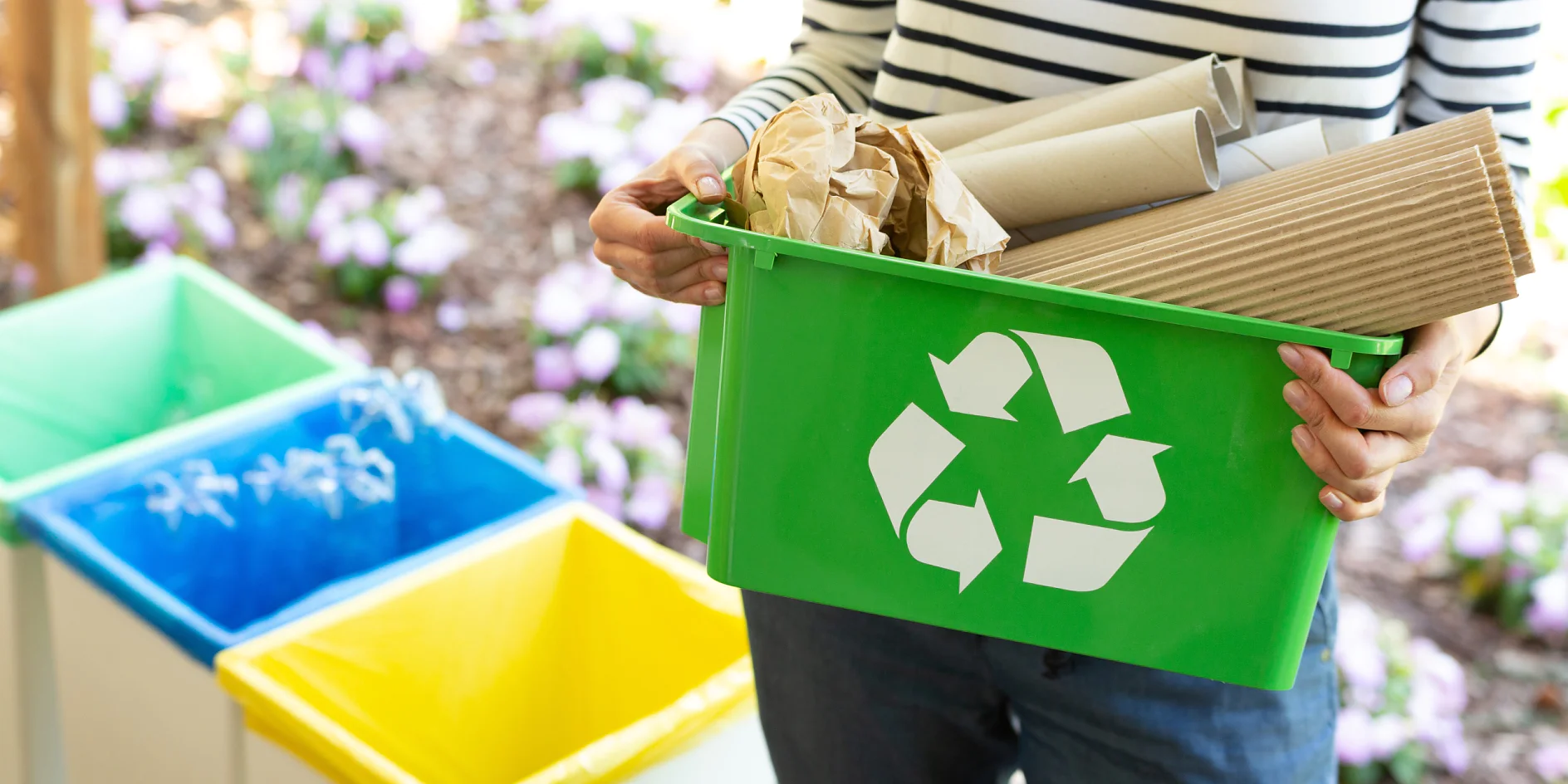 A person holds a green recycling bin filled with brown cardboard and paper secured with tesa tape. The bin features a white recycling symbol. In the background, there are blue, yellow, and green bins, along with flowerbeds showcasing purple and white flowers. (This text has been generated by AI)