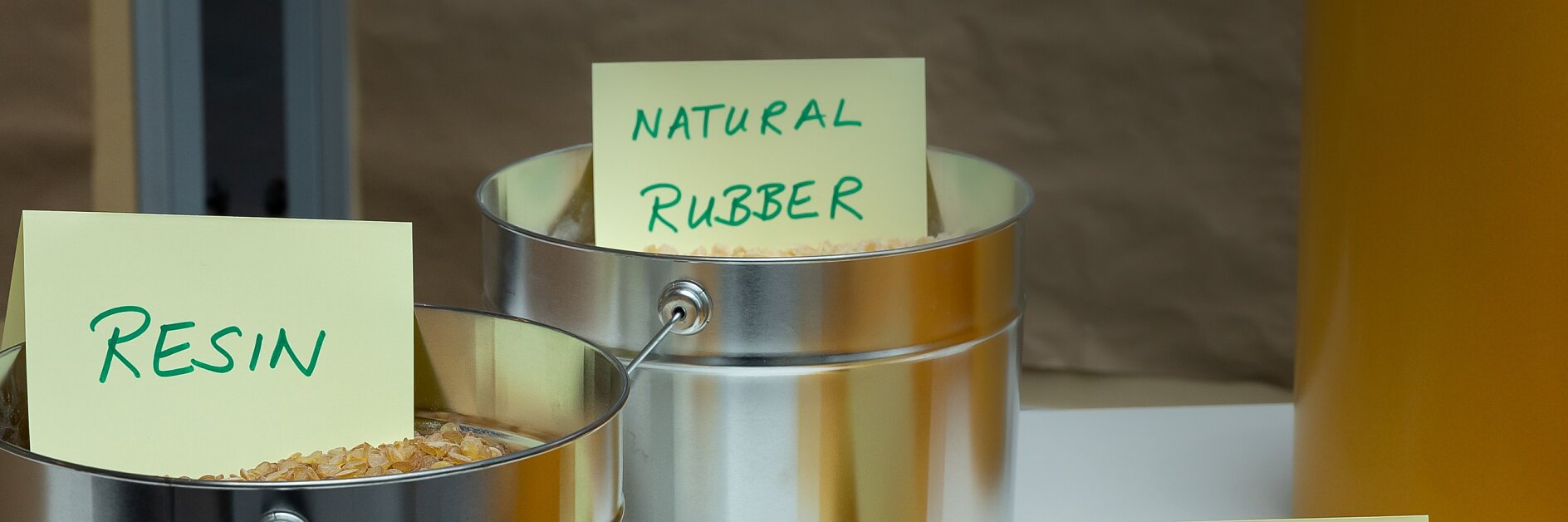 Metal containers labeled Resin and Natural Rubber hold raw materials. A tesa tape Liner sign is placed in front of yellow rolls on a table. (This text has been generated by AI)