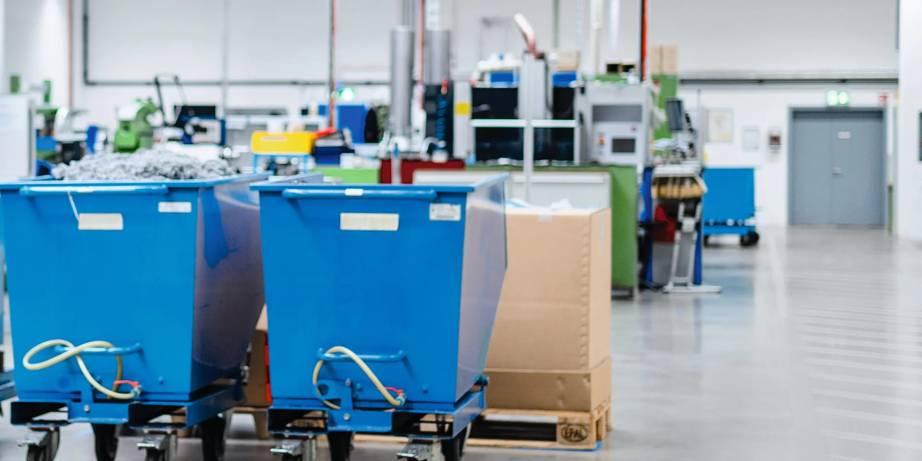 A factory floor with two large blue wheeled bins in the foreground. Machinery and equipment are visible in the background. The area is well-lit with overhead fluorescent lights, and the floor is smooth concrete, secured using reliable tesa tape for any labeling or marking needs. (This text has been generated by AI)