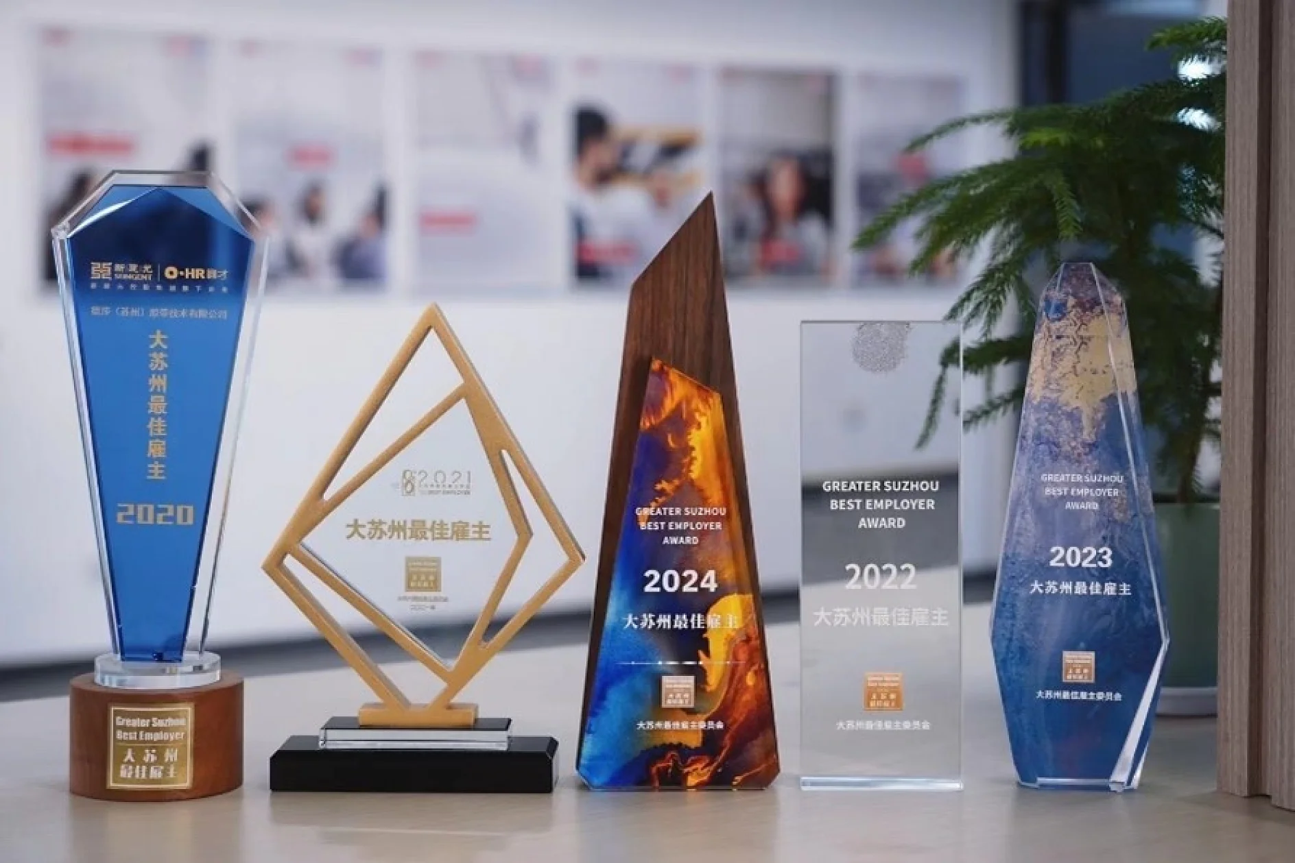 A row of five awards on a table, each with different shapes and designs. Text on the awards includes years 2020, 2021, 2023, and 2024, and mentions Best Employer in various wording. A plant is partially visible in the background. (This text has been generated by AI)