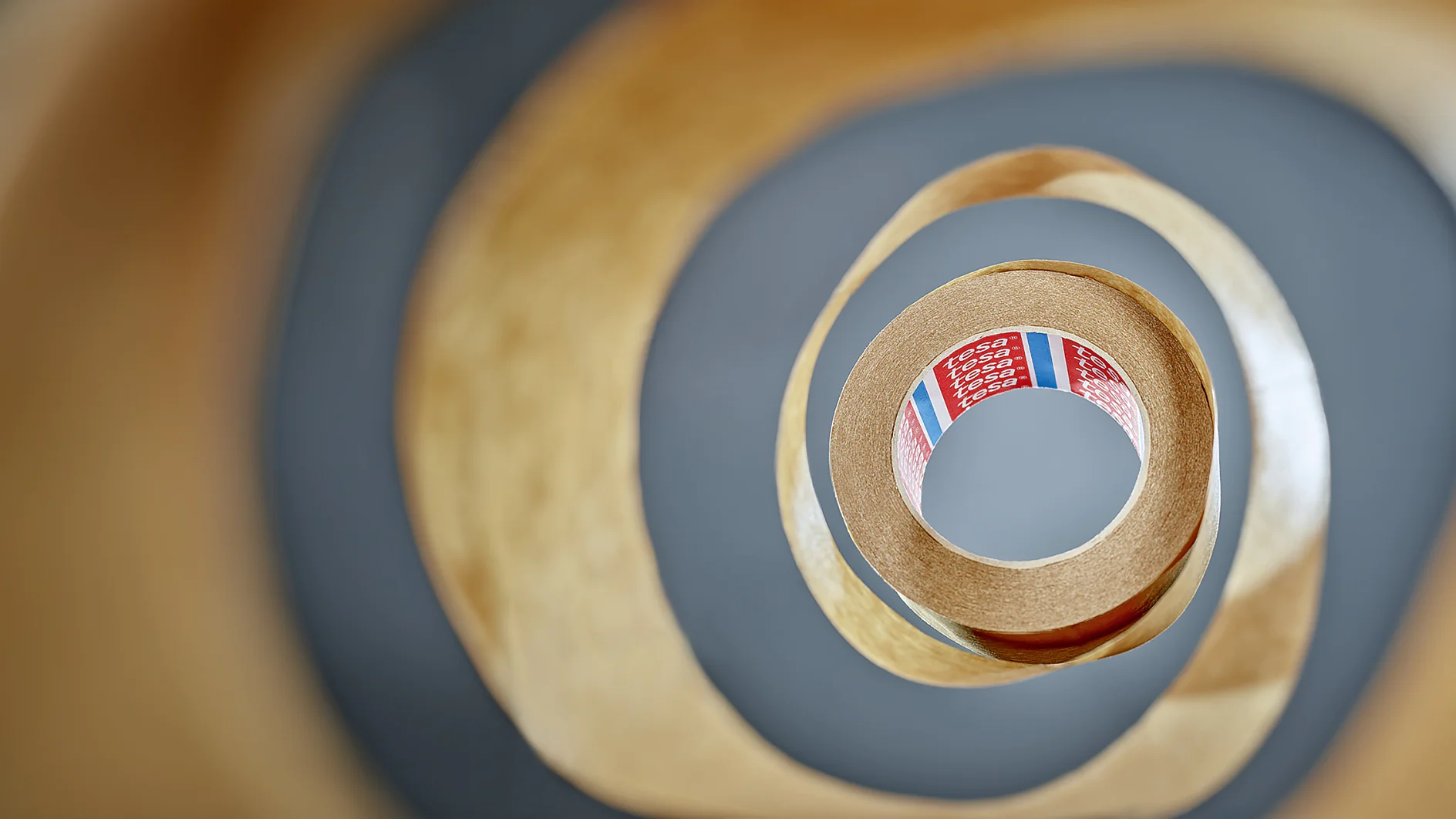 View from the center of a roll of tesa tape, creating a spiral pattern. The tesa tape is brown with a partially visible red label inside, featuring text and colored stripes. The background is blurred, emphasizing the layered effect of the tesa tape. (This text has been generated by AI)